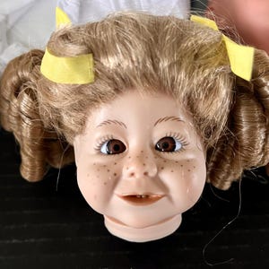 May include: A porcelain doll head with brown curly hair and yellow hair bows. The doll has blue eyes, rosy cheeks, and a freckled nose. The doll is smiling.