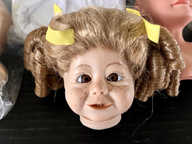 May include: A porcelain doll head with brown curly hair and yellow hair bows. The doll has blue eyes, rosy cheeks, and a freckled nose. The doll is smiling.