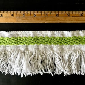 Op de afbeelding: Witte bies met een groene geweven band en lange witte franjes. De bies wordt gemeten met een houten liniaal met inchmarkeringen.