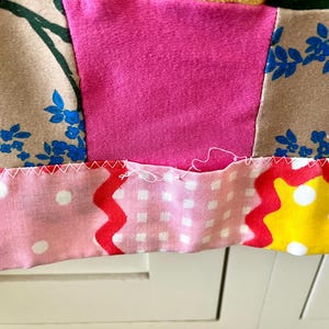 May include: A patchwork textile featuring a variety of fabric patterns including beige with blue floral prints, solid hot pink, light pink with white polka dots, and yellow with white polka dots, joined by white zigzag stitching.