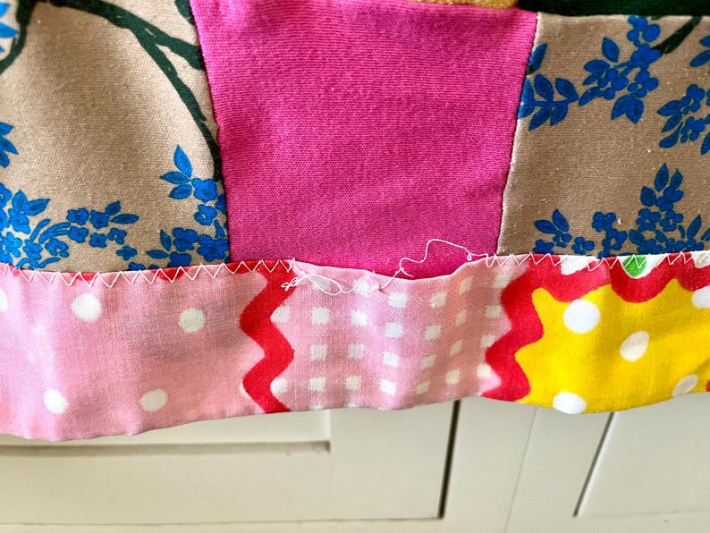May include: A patchwork textile featuring a variety of fabric patterns including beige with blue floral prints, solid hot pink, light pink with white polka dots, and yellow with white polka dots, joined by white zigzag stitching.