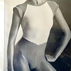 May include: A vintage leotard in black and white. The leotard features a white bodice with short, sheer sleeves and a scoop neckline. The lower portion is a darker color, with a high-cut leg design. The leotard is likely for dance or gymnastics.