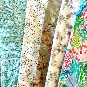May include: A collection of folded fabrics with various patterns. Designs include a gray and white plaid, teal floral, confetti dots, floral with roses, and a vibrant tropical print with a mermaid.