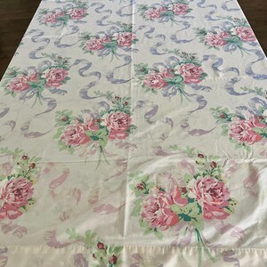 May include: A white tablecloth with a repeating pattern of pink roses, green leaves, and lavender ribbons. The floral design is evenly spaced across the fabric, creating a vintage aesthetic. The tablecloth is spread out on a table.