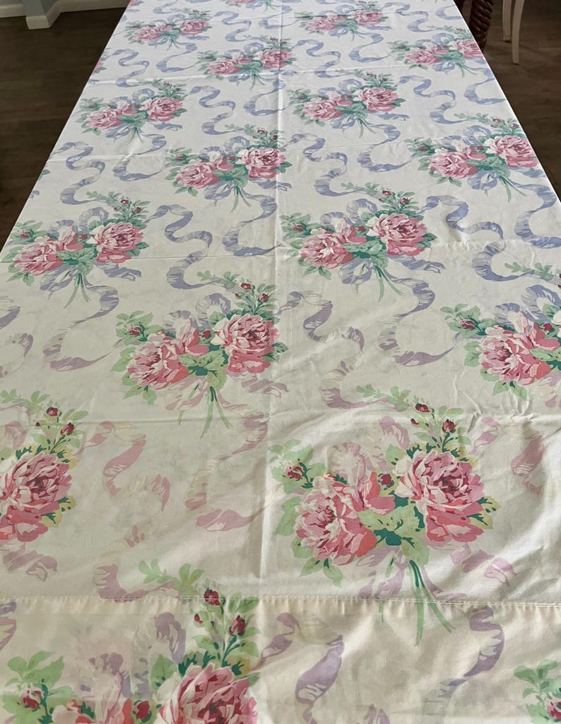 May include: A white tablecloth with a repeating pattern of pink roses, green leaves, and lavender ribbons. The floral design is evenly spaced across the fabric, creating a vintage aesthetic. The tablecloth is spread out on a table.