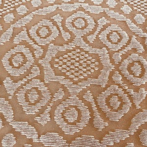 May include: Close-up of a tan textile with a repeating pattern of white, stylized floral and geometric shapes. The design features a central medallion with radiating elements and a textured appearance, suitable for home decor.