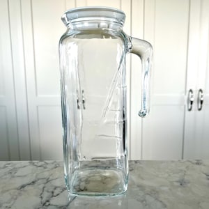 May include: A clear glass pitcher with a rectangular shape and a handle. The pitcher has a lid and appears to be empty. The pitcher is sitting on a marble countertop in front of a white cabinet.