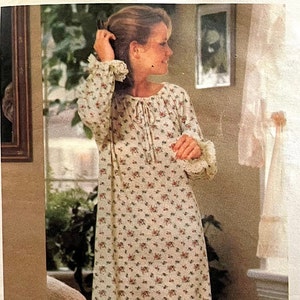 Op de afbeelding: Een witte nachtjapon met een bloemenprint en kantversiering. De nachtjapon heeft een strik aan de hals en lange mouwen.