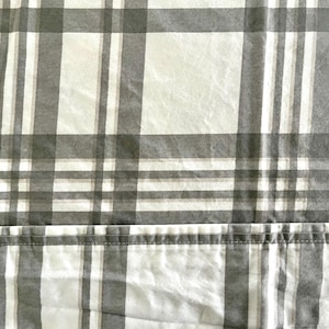 May include: Close-up of a fabric with a plaid pattern. The design features a grid of gray and white lines, creating a checkered effect. The fabric appears to be a light material, possibly cotton or linen, suitable for bedding or apparel.