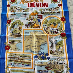May include: A colorful tea towel with a map of Devon, England, and illustrations of various towns and landmarks. The text "Memories of Devon" is printed in the center of the towel. The towel is bordered with red and white flowers.