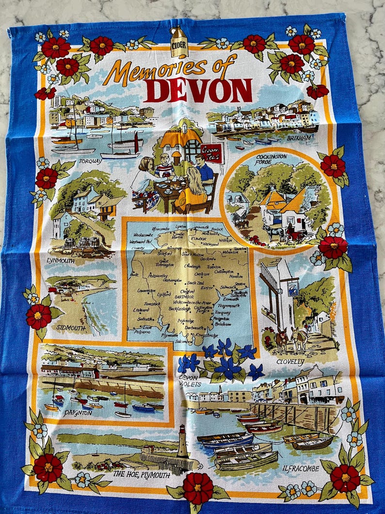May include: A colorful tea towel with a map of Devon, England, and illustrations of various towns and landmarks. The text "Memories of Devon" is printed in the center of the towel. The towel is bordered with red and white flowers.