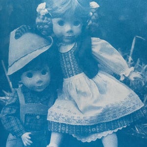 May include: A blue-toned monochromatic print featuring two dolls. One doll wears a hat and plaid shirt with overalls, while the other doll has braided hair with bows, wearing a dress with a patterned bodice, a white apron, and lace trim.