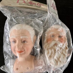 May include: Two porcelain doll heads in a plastic bag. The heads are of an older woman and Santa Claus. The woman's head has white hair and a smiling face. Santa's head has a white beard and a rosy complexion. The package says "Porcelain Mrs. Santa & Santa".