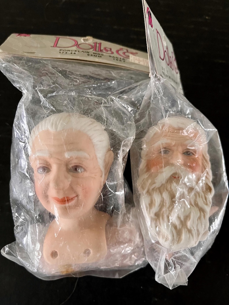 May include: Two porcelain doll heads in a plastic bag. The heads are of an older woman and Santa Claus. The woman's head has white hair and a smiling face. Santa's head has a white beard and a rosy complexion. The package says "Porcelain Mrs. Santa & Santa".