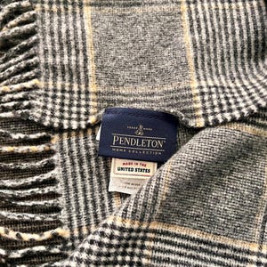 May include: A grey, black, and cream plaid throw blanket with fringe edges. A dark blue Pendleton Home Collection label is stitched to the fabric, featuring the text Trade Mark, Pendleton, Home Collection, and Made in the United States.