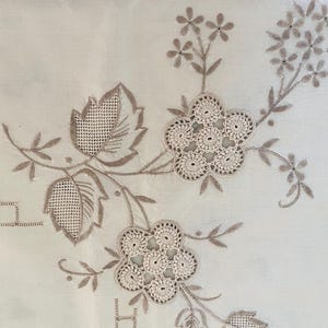 May include: A white fabric textile featuring intricate beige embroidery. The design includes two large floral motifs with openwork centers, detailed leaves with cross-hatched stitching, and delicate stems with small flower buds and geometric borders.