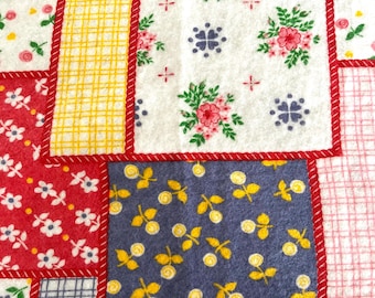 Op de afbeelding: Een patchwork stof met verschillende bloem- en geometrische patronen in rood, geel, blauw en wit. De stof is met rode stiksels aan elkaar genaaid.