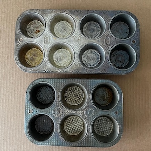 May include: Two vintage metal muffin tins. The tins are rectangular with rounded cups, each with a textured, circular pattern at the bottom. The metal has a worn, aged appearance, with some discoloration and signs of use. Ideal for baking.