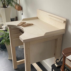 Könnte beinhalten: Eine helle Werkbank für Juweliere aus Holz mit einer geschwungenen Aussparung an der Vorderkante der Tischplatte, einer erhöhten Rückwand und einem kleinen hölzernen Feilnagel auf der Oberfläche. Die Konstruktion hat vier stabile Beine und Ablagen.