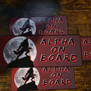 May include: Rectangular stickers with a red background and the words "ALPHA ON BOARD" in white with a black outline. Each sticker features a silhouette of a wolf howling at the moon. The stickers are arranged on a wooden surface.
