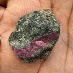 May include: A raw, unpolished mineral specimen featuring a dark, textured grey-green matrix stone with a prominent, embedded band of translucent, deep magenta-pink crystalline material running diagonally across the center of the rough rock.