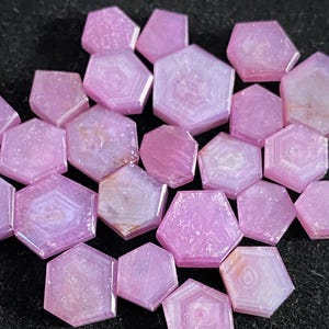 Puede incluir: Un conjunto de gemas hexagonales rosas con diversas tonalidades y diseños internos. Las gemas están dispuestas sobre una superficie oscura y texturizada, mostrando sus facetas pulidas y su calidad translúcida. Las piedras son de color rosa.