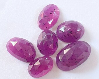 Può includere: Una collezione di sei gemme color magenta di forma ovale e sfaccettate. Ogni pietra presenta una superficie lucida con un taglio geometrico e inclusioni scure visibili sparse nel materiale rosa-viola traslucido delle gemme.