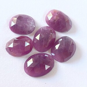 Peut inclure: Six pierres précieuses ovales et facettées, dans des tons de violet et de rose, sont disposées sur une surface blanche. Les pierres ont une surface polie et réfléchissante, avec des facettes visibles qui captent la lumière. L'esthétique générale est celle de l'élégance et de la beauté naturelle.