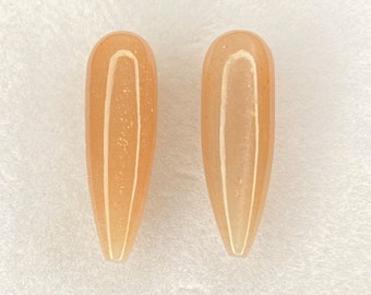 May include: Two almond-shaped, light orange-coloured nail tips with a glossy finish. The tips are displayed on a white surface inside a clear plastic container. The nails have a subtle shimmer.