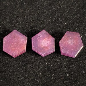 May include: Three translucent, hexagonal-shaped objects in a vibrant magenta-pink hue. Each piece features a smooth, flat surface with a subtle, internal geometric pattern visible through the semi-transparent material, arranged in a horizontal row.