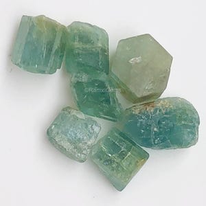 Puede incluir: Un grupo de seis cristales de aguamarina verde de diferentes formas y tamaños. Los cristales son translúcidos y tienen un color verde claro.