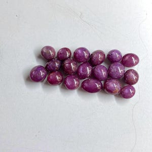 May include: A collection of polished, oval and round gemstones in shades of deep purple and burgundy. The stones have a smooth, reflective surface, and are arranged on a white background. The gemstones vary in size and shape.