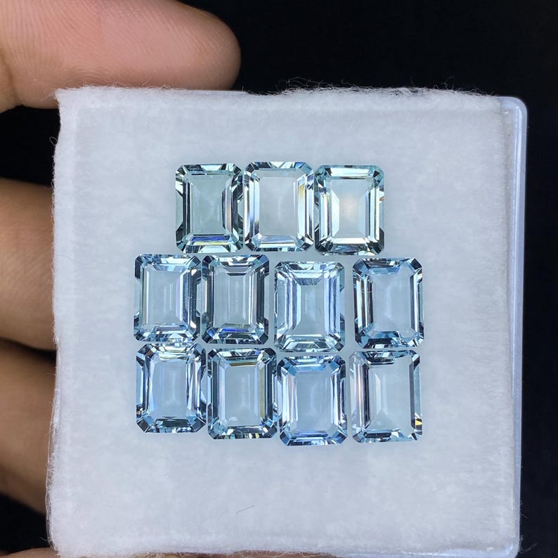 Puede incluir: Una colecci&oacute;n de piedras preciosas de aguamarina azul claro en talla esmeralda cuadrada. Las piedras preciosas est&aacute;n dispuestas en forma de pir&aacute;mide en una caja blanca.