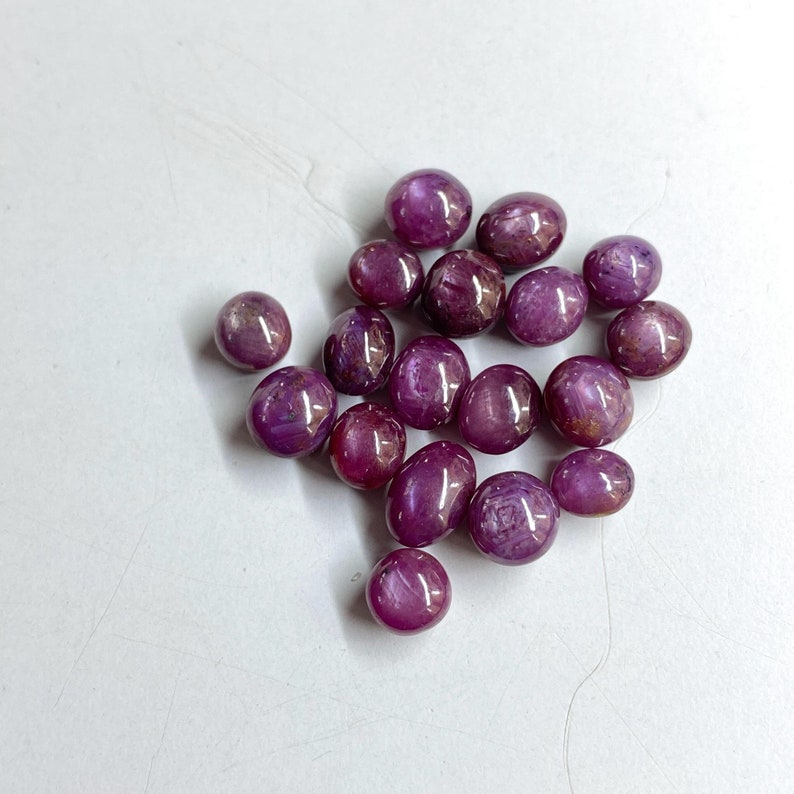 May include: A collection of polished, deep red gemstones with a glossy finish. The stones vary in size and shape, with a mix of rounded and irregular forms. The gems are scattered on a white surface, creating a contrast of color and texture.