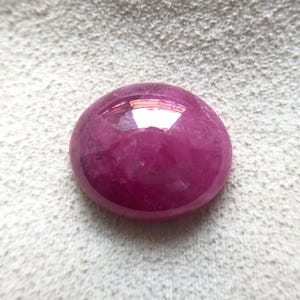 Può includere: Una gemma di rubino lucidata, taglio cabochon. La gemma è di un colore rosso-rosato intenso e saturo, con una superficie liscia e arrotondata. La pietra riflette la luce.