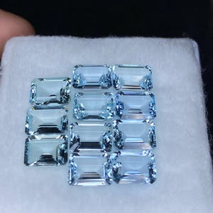 Puede incluir: Una colecci&oacute;n de doce piedras preciosas de aguamarina azul claro, talladas en forma de esmeralda, expuestas sobre una superficie blanca.