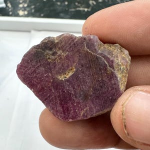 Peut inclure: Une pierre précieuse brute et non polie aux teintes magenta profond et violet foncé. La pierre présente une surface rugueuse et texturée avec des stries visibles, des inclusions minérales naturelles et des bords irréguliers, d'aspect translucide.