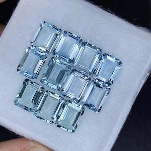 Puede incluir: Una colecci&oacute;n de doce piedras preciosas de aguamarina azul claro, talladas en forma de esmeralda rectangular, expuestas sobre una superficie blanca.