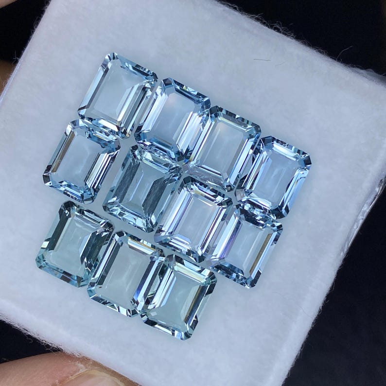 Puede incluir: Una colecci&oacute;n de doce piedras preciosas de aguamarina azul claro, talladas en forma de esmeralda rectangular, expuestas sobre una superficie blanca.