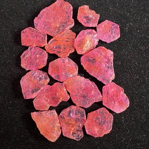 May include: A collection of pink ruby rough gemstones. The rough gemstones are various shapes and sizes. The gemstones are on a black background.