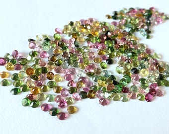 Puede incluir: Una colección de piedras preciosas redondas y facetadas en varios tonos de verde, rosa y amarillo. Las piedras preciosas están dispersas sobre una superficie blanca.