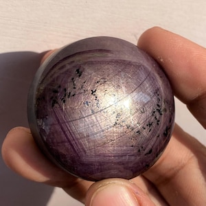 Puede incluir: Una gema esférica pulida, en tonos morados y grises, sostenida en una mano. La superficie tiene una cualidad reflectante con vetas e inclusiones oscuras. La esfera tiene aproximadamente 5 cm de diámetro.