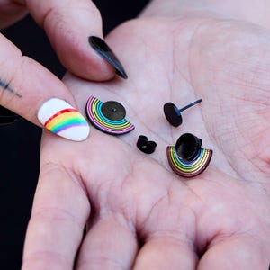 Peut inclure: Une paire de boucles d'oreilles à clous arc-en-ciel avec des tiges et des fermoirs noirs. Les boucles d'oreilles ont la forme d'un demi-cercle avec un motif arc-en-ciel. Elles sont tenues dans une main avec un ongle peint en arc-en-ciel.