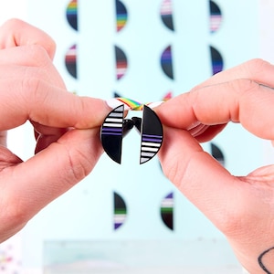 May include: Black and white half-circle earrings with purple and white stripes. The earrings are held in front of a light blue background with other half-circle designs in various colors. The earrings have a post backing.