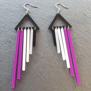 May include: A pair of geometric earrings featuring black triangle tops with silver hooks. Each earring has a series of dangling, vertical bars in purple, silver, and white. The earrings are designed to be lightweight and eye-catching.