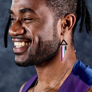 May include: A person wearing geometric dangle earrings. The earrings have a black triangle top with vertical bars in purple, white, and teal. The person is wearing a purple top and a silver chain necklace.