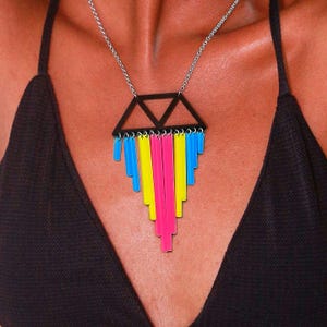 May include: A geometric pendant necklace with a black triangle and a silver chain. The pendant features vertical bars in blue, yellow, and pink. The necklace is worn against a dark background.