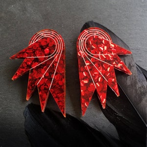 May include: A pair of red glitter earrings with a geometric design. The earrings are shaped like a stylised sunburst with white lines.
