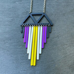 Könnte beinhalten: Halskette mit silberner Kette und einem schwarzen geometrischen Anhänger. Der Anhänger hat ein Dreieck-Design und vertikale Stäbe in Lila, Weiß, Gelb und Schwarz, was einen farbenfrohen, modernen Look erzeugt.