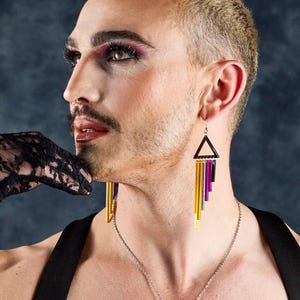 Op de afbeelding: Een persoon die geometrische oorbellen draagt met een zwarte driehoekige top en verticale gouden, paarse en zilveren staven. De persoon draagt een zwarte top en een zilveren ketting. Een zwarte kanten handschoen bevindt zich in de buurt van het gezicht.